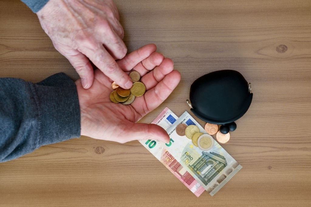 Mãos segurando moedas douradas em uma mesa com cédulas de dinheiro, moedas e uma pequena carteira ao lado.