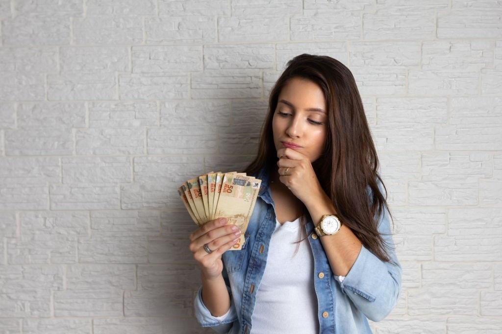 Uma mulher pensando enquanto segura dinheiro na mão.